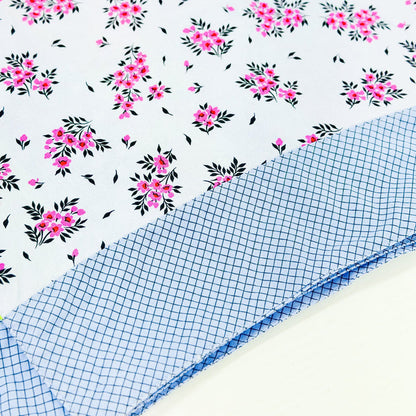 A folded piece of fabric with a white background printed with pink floral clusters and black leaves, layered over a folded piece of light blue and white gingham fabric.