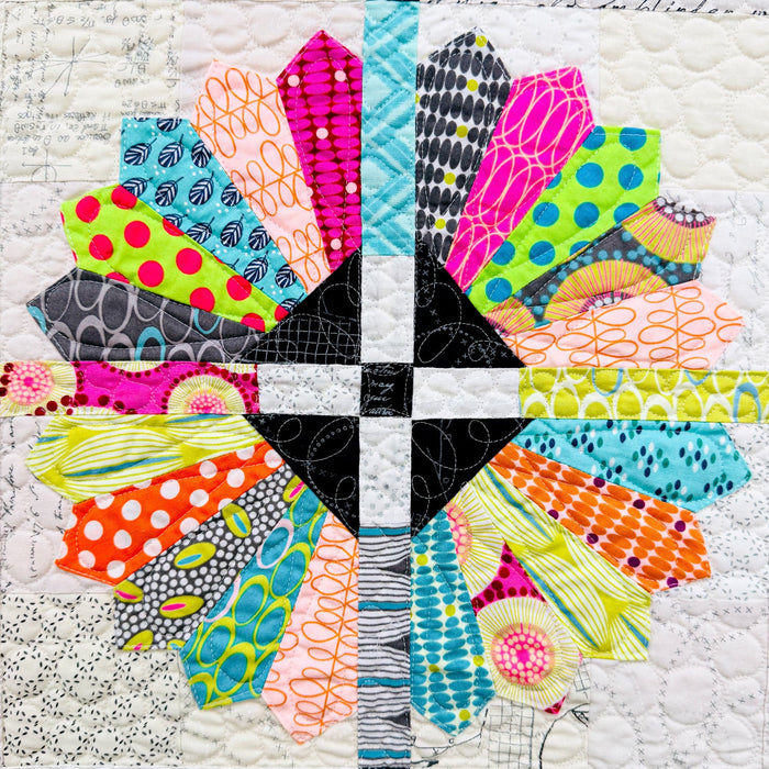 A close-up of a colorful quilted fabric design featuring a symmetrical cross shape made of multiple patterned fabric sections in bright neon, orange, pink, yellow, blue, green, black, and white.