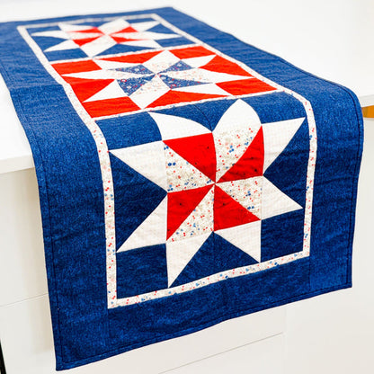 A rectangular quilted table runner with a red, white, and blue patriotic starburst pattern draped over a white table against a white wall.
