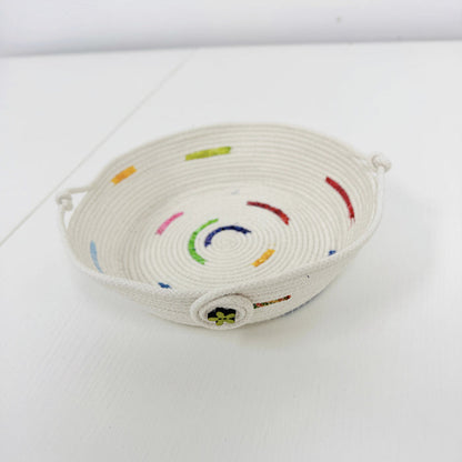A small woven basket tray made from off-white rope, decorated with colorful embroidered curved lines and small dots in red, green, blue, yellow, and pink, set against a plain white background.