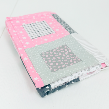 A folded handmade quilted baby blanket with a house design, featuring pink starry fabric, a gray floral patch, and striped details, displayed on a white surface.